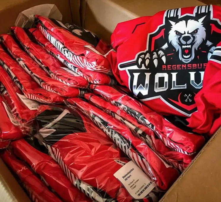 Une boîte en carton contenant plusieurs maillots de sport rouges personnalisés emballés dans du plastique, l'un d'eux affichant un logo de loup et l'inscription 'REGENSBURG WOLVES'.