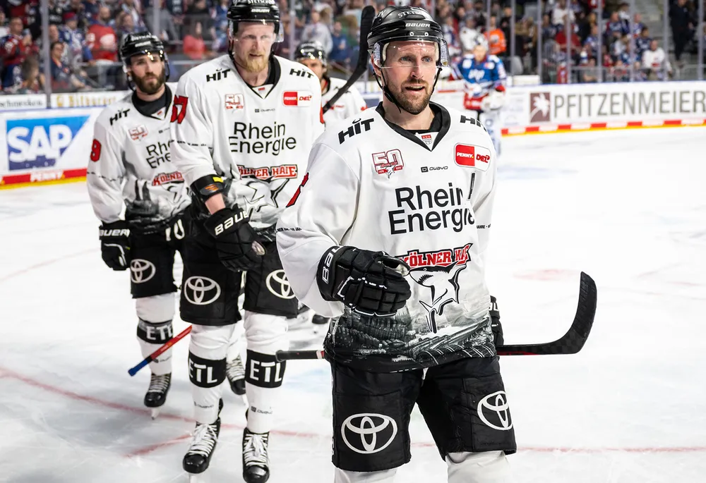 Joueurs de hockey sur glace en maillots blancs avec des pantalons noirs portant le logo d'une équipe, debout sur la glace.