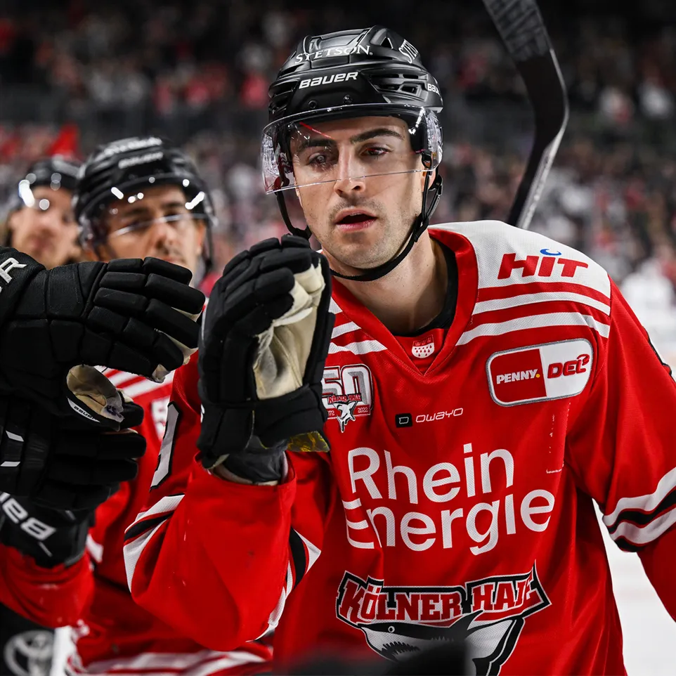 Un joueur de hockey dans un maillot rouge avec des rayures blanches et des logos, levant sa main en célébration.