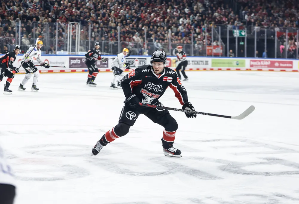 Un joueur de hockey sur glace portant un maillot noir avec des accents rouges et blancs, patinant sur la glace avec une crosse.