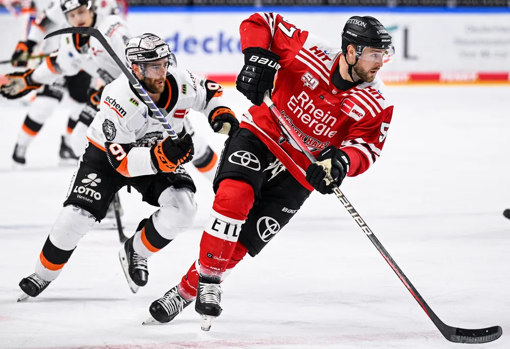 Un joueur de hockey en maillot rouge patine avec le palet sur la glace pendant qu'un autre joueur en maillot blanc le suit de près.