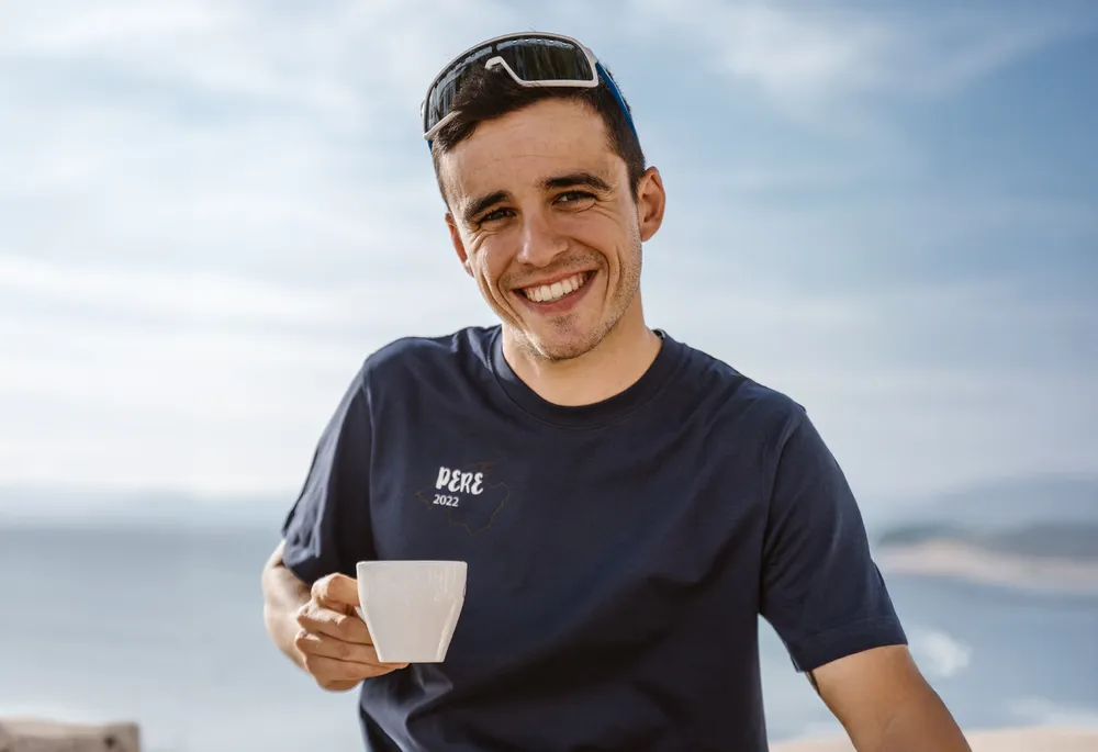 Un homme souriant portant un t-shirt bleu tient une tasse à la main, sur fond d'un magnifique paysage côtier.