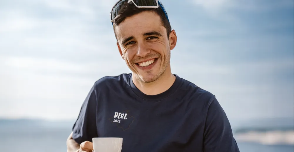 Un homme sportif portant un t-shirt bleu marine avec l'impression 'PERE 2022' sourit en tenant une tasse, devant un arrière-plan côtier flou.