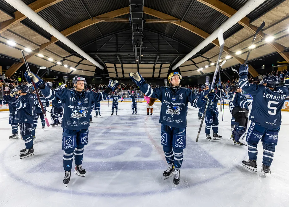 Des joueurs de hockey sur glace en maillots bleus se tiennent sur la glace, célébrant avec des bâtons levés devant une foule en délire.