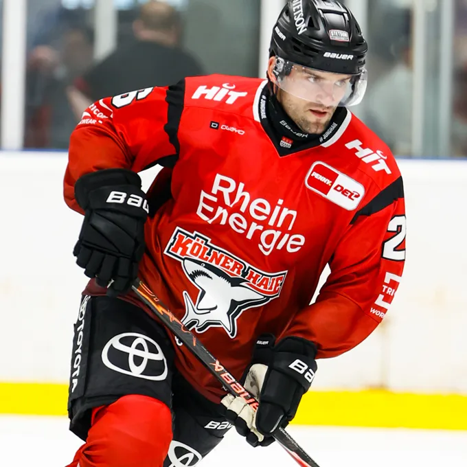Un joueur de hockey sur glace portant un maillot rouge avec le logo de Rhein Energie et Kölner Haie, sur la glace.