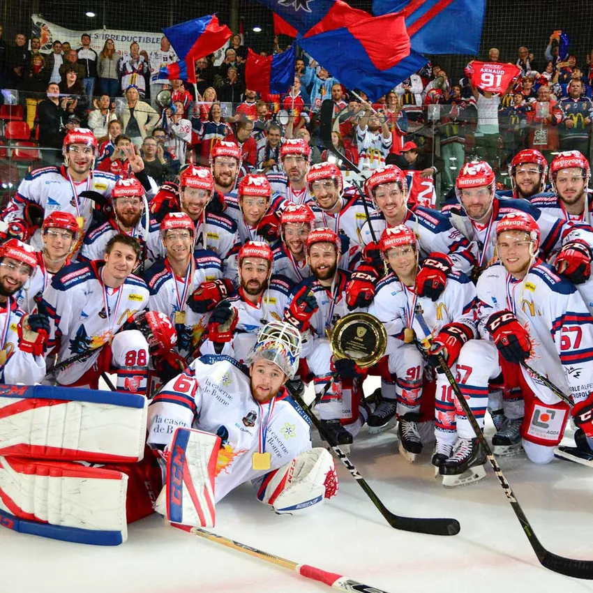 Un groupe de joueurs de hockey sur glace en maillots blancs avec des accents rouges et bleus pose fièrement avec des médailles et un trophée sur la glace.