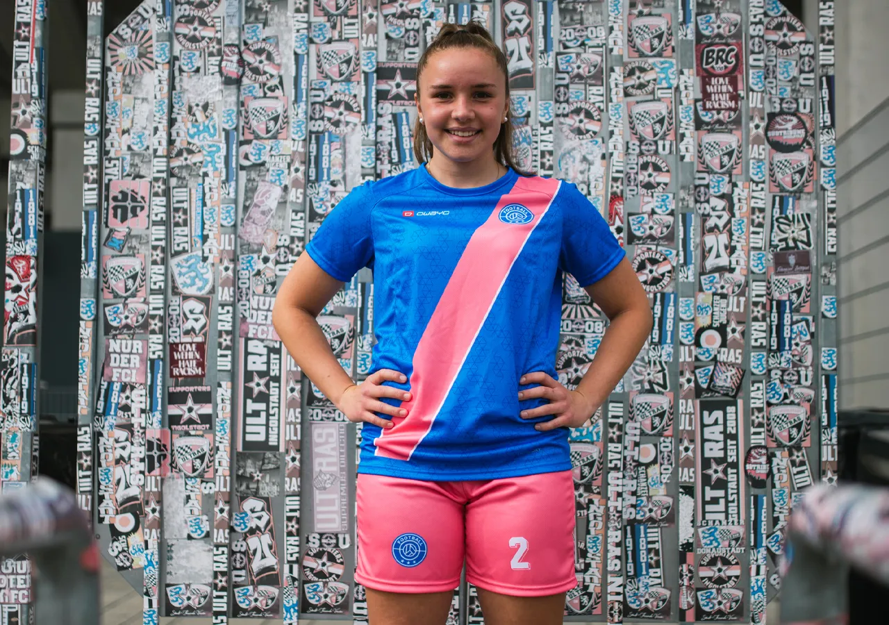 Une jeune femme porte un maillot de football bleu avec une bande diagonale rose et un short rose avec le numéro 2, devant un mur recouvert de graphismes et d'autocollants.