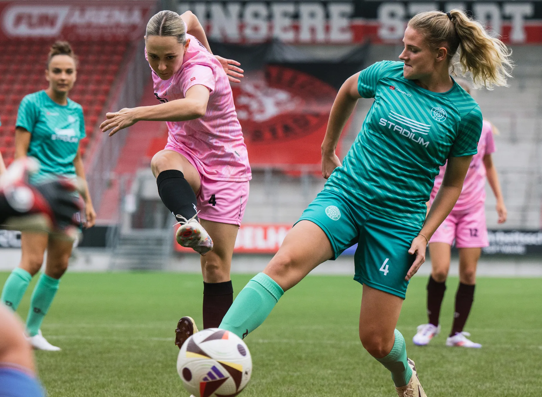 Deux footballeuses en maillots colorés, l'une en rose et l'autre en vert, jouent sur un terrain en herbe.