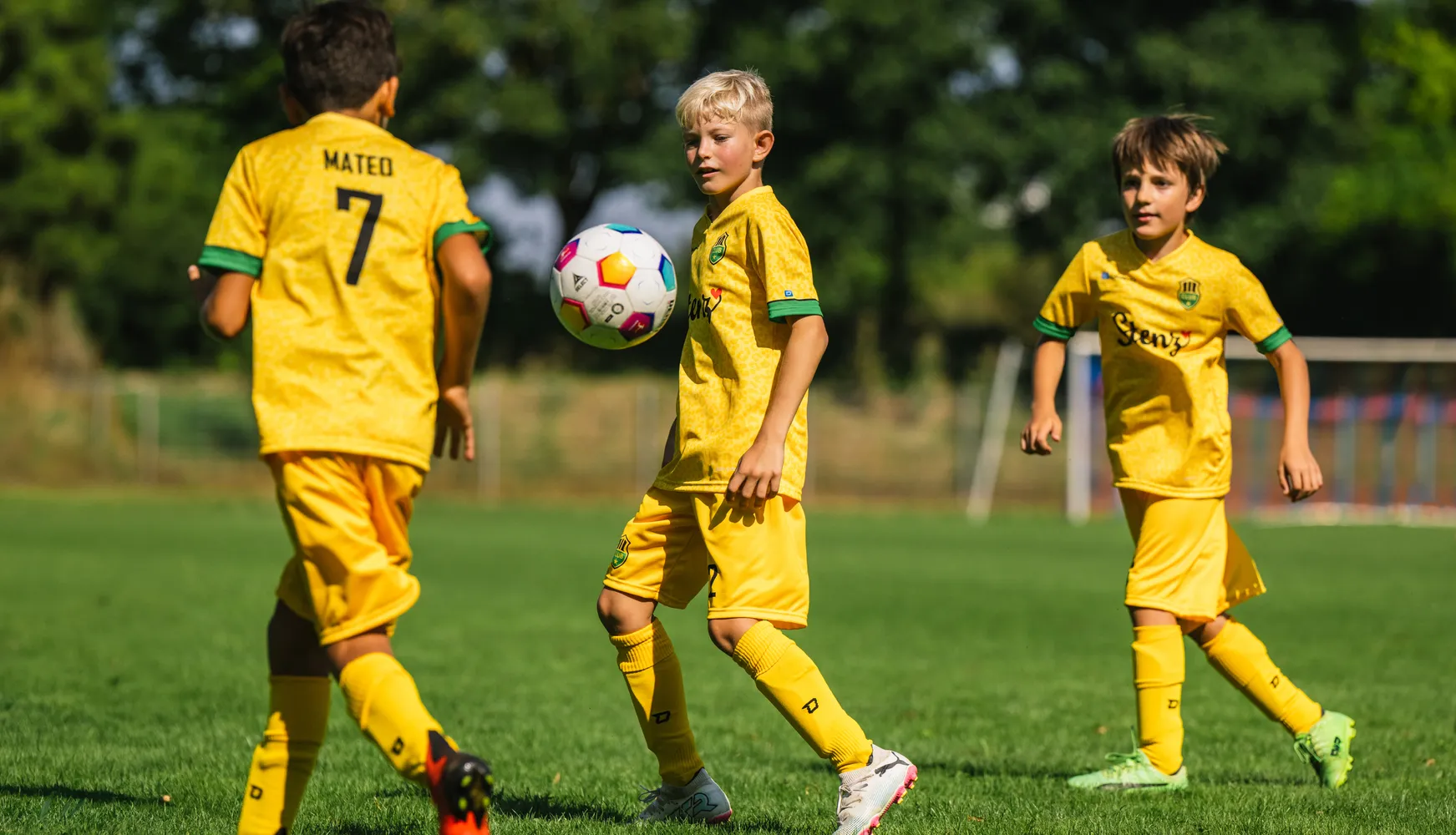 Trois enfants en maillots de football jaunes jouent sur un terrain gazonné avec un ballon de football coloré.