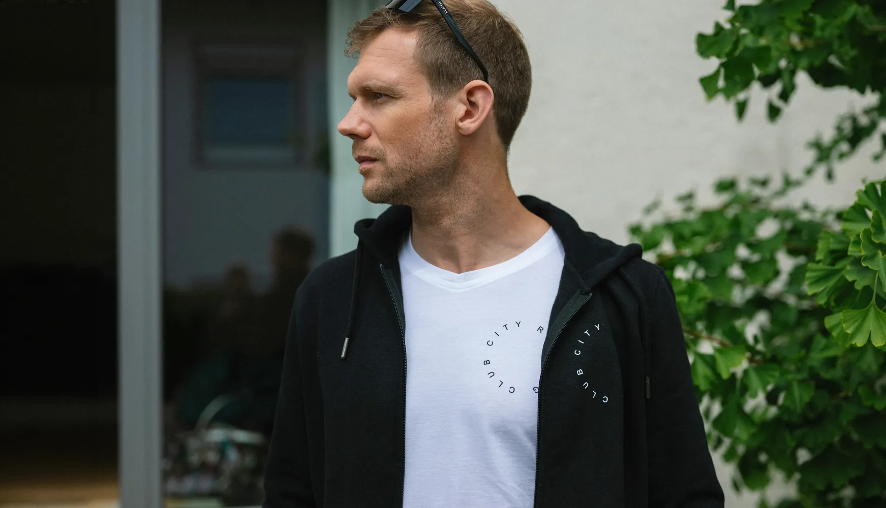 Un homme portant un sweat à capuche noir sur un t-shirt blanc avec un imprimé circulaire se tient dans un espace extérieur.