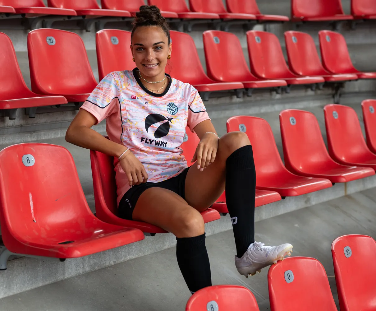 Une jeune femme est assise sur un siège de stade rouge, portant un maillot de football coloré et des chaussettes noires.