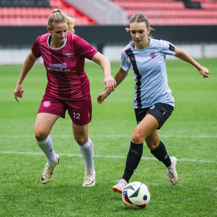 Deux femmes en maillots de football jouent sur un terrain en herbe, tentant de contrôler le ballon.