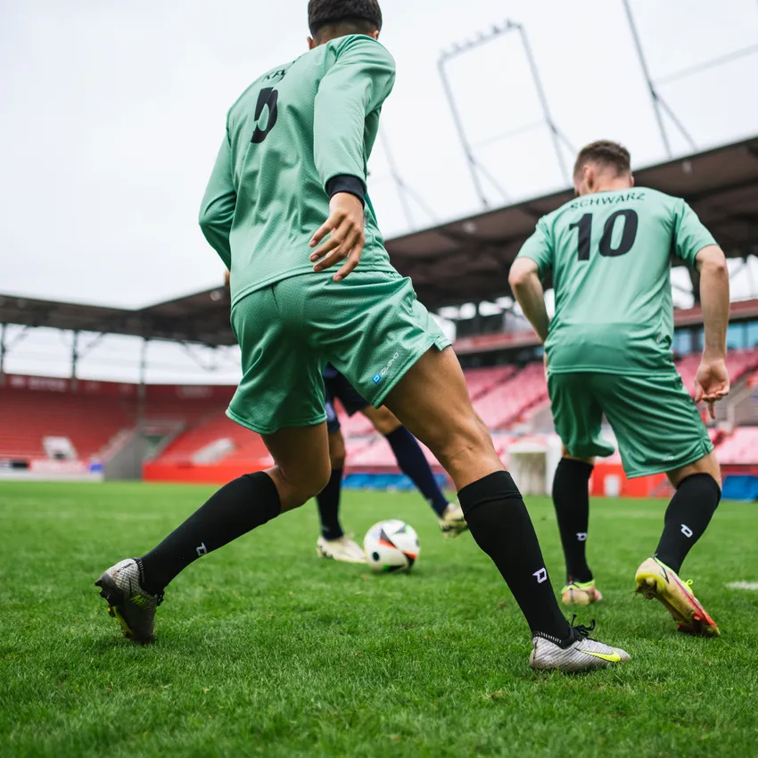 Deux joueurs en maillots de football verts et chaussettes noires jouant sur un terrain dans un stade.