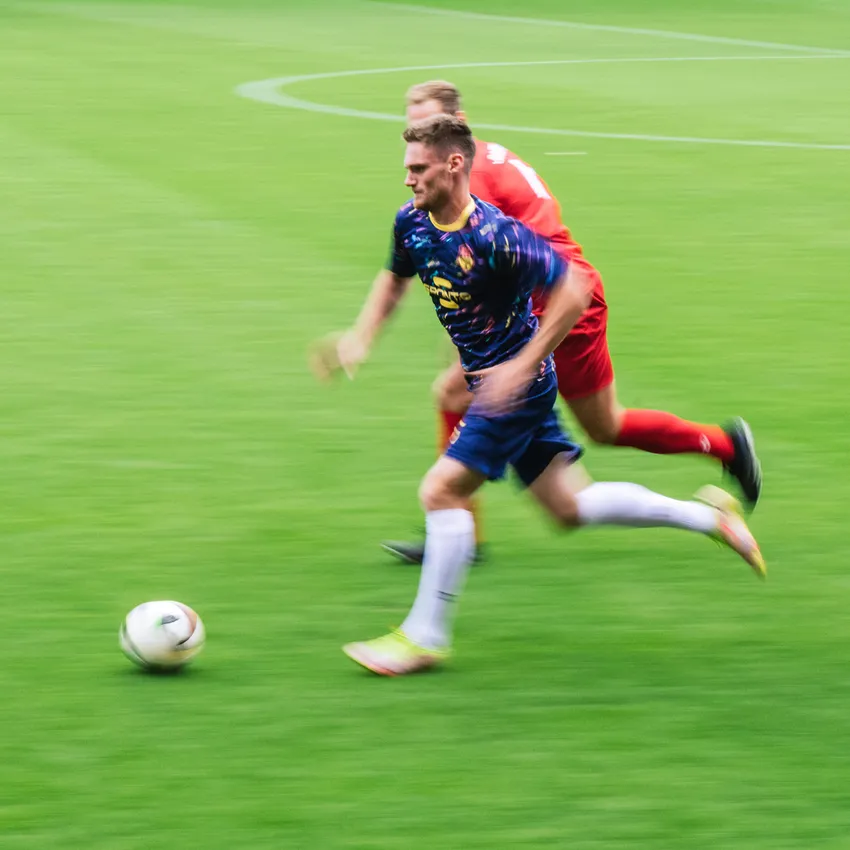 Un joueur de football en maillot coloré sprinte sur le terrain vert tandis qu'un autre joueur le poursuit.