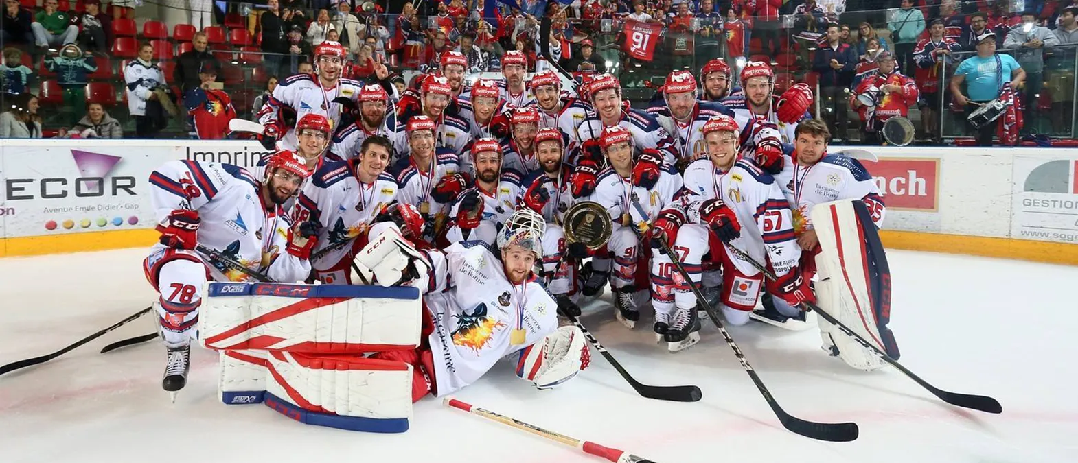Une équipe de hockey sur glace en maillots blancs avec des accents rouges et bleus pose sur la glace, entourée de supporters, tandis que les joueurs portent des médailles et tiennent un trophée.
