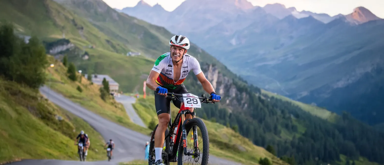 Un cycliste en maillot blanc, vert et orange avec le numéro 29 roule sur une route de montagne dans un paysage montagneux.