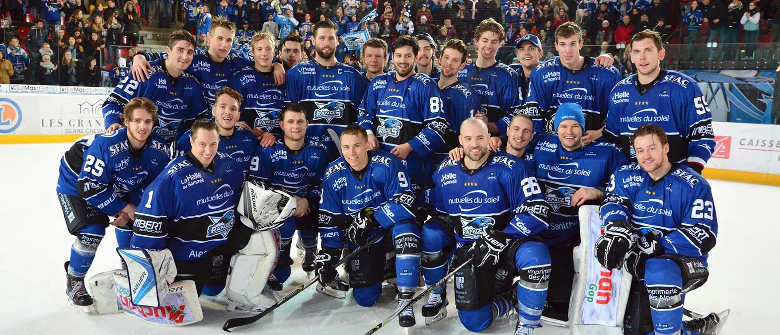 Une équipe de hockey sur glace en maillots bleus avec le logo des Rapaces de Gap pose sur la glace devant un public.