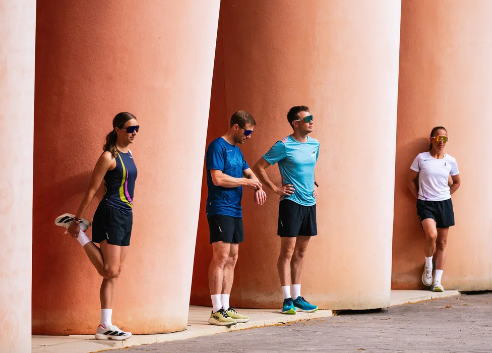 Un groupe de quatre athlètes en tenue de course s'étire contre un mur avec de grandes colonnes.