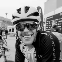 Un cycliste souriant portant une veste de cyclisme noire avec l'inscription 'BIKE INDUSTRIES' sur la manche et un casque blanc, portant des lunettes de soleil reflétant l'environnement.