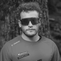 Un homme aux cheveux bouclés et aux lunettes de soleil porte un t-shirt de sport gris à manches longues avec le logo 'owayo' sur l'épaule et d'autres logos de sponsors sur la poitrine.