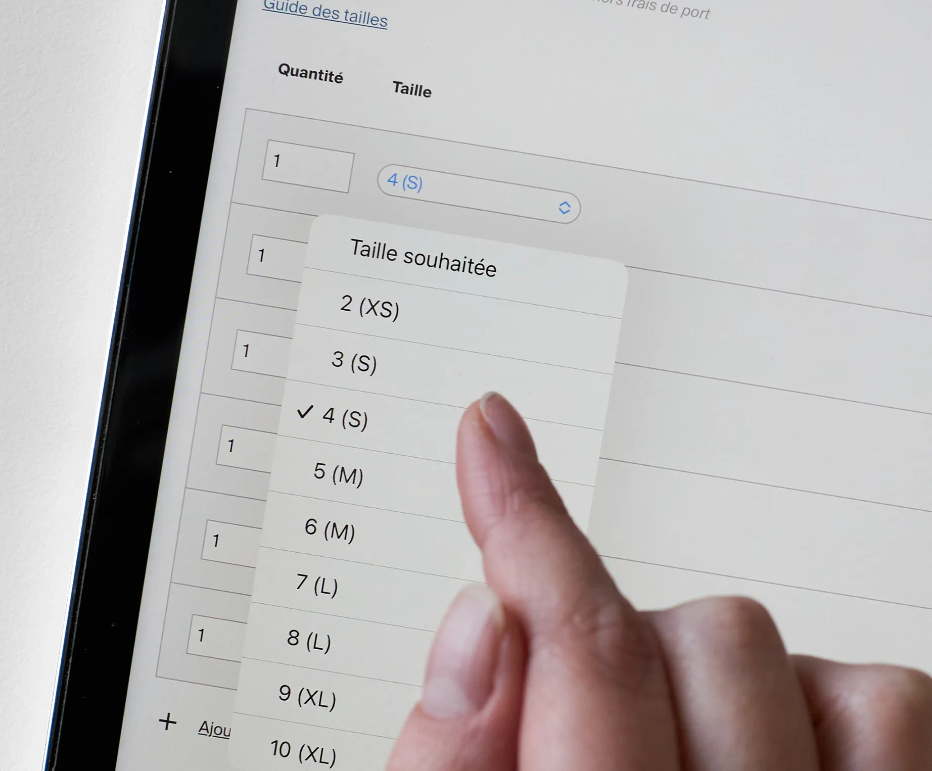Une main sélectionne la taille 4 (S) dans une liste sur un écran de tablette affichant un guide des tailles pour des vêtements de sport personnalisés.