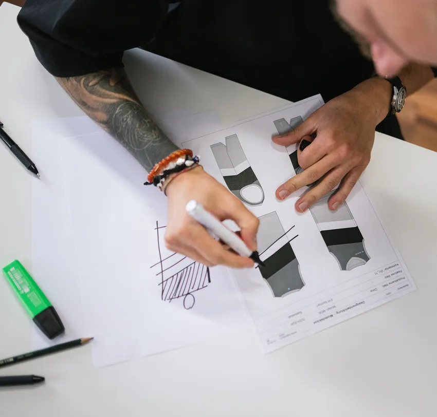 Une personne avec un bras tatoué dessine des modèles pour des vêtements de sport personnalisés avec un marqueur sur une feuille de papier.