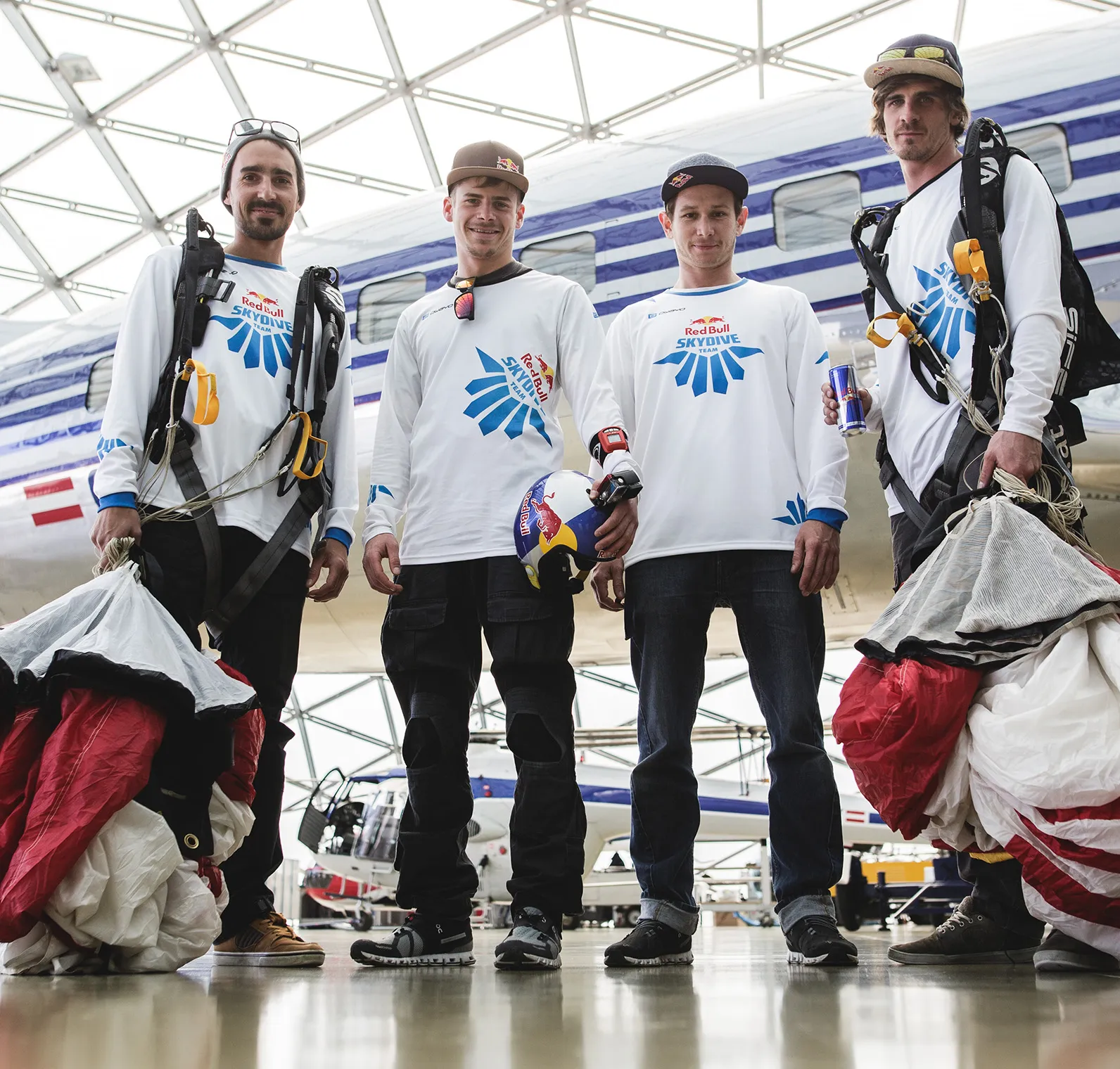 Quatre hommes portant des t-shirts blancs à manches longues avec le logo de l'équipe Red Bull Skydive se tiennent dans un hangar devant un avion.