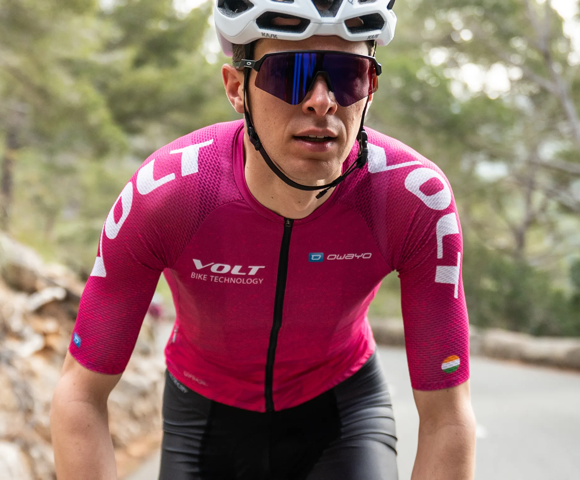 Un cycliste portant un maillot de cyclisme rose à manches courtes avec le logo 'VOLT BIKE TECHNOLOGY' et le logo 'owayo' sur la poitrine, roulant sur une route dans une zone boisée.