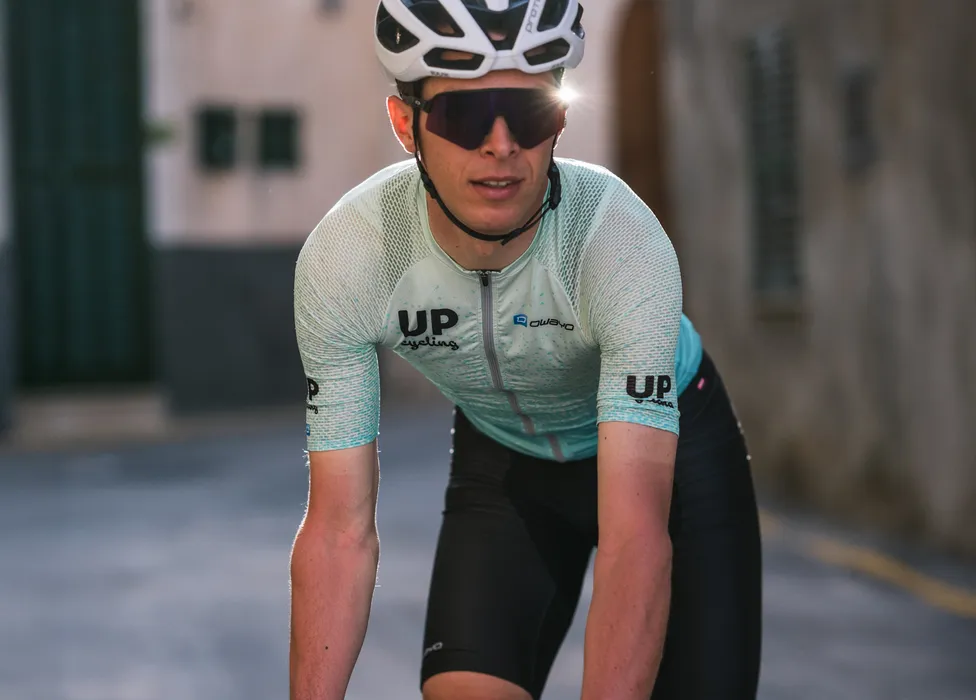 Un cycliste portant un maillot de cyclisme bleu clair respirant avec les logos 'UP cycling' et 'owayo' sur la poitrine, dans une posture sportive sur une rue.