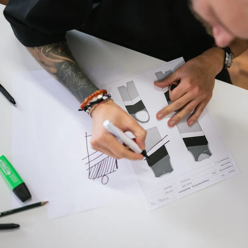 Une personne avec un bras tatoué esquisse des designs de vêtements de sport sur du papier, entourée d'outils d'écriture.