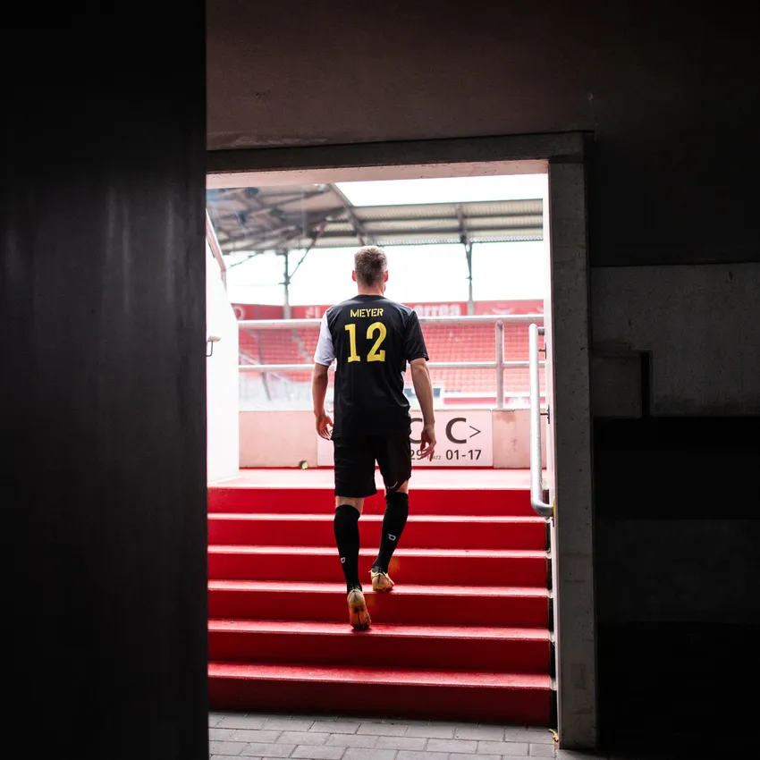 Un footballeur en maillot noir avec le numéro 12 monte les marches rouges d'un stade.