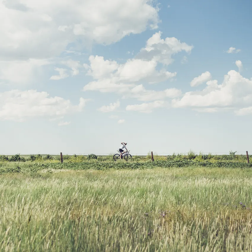 Une personne fait du vélo à travers un paysage vert sous un ciel bleu avec quelques nuages.