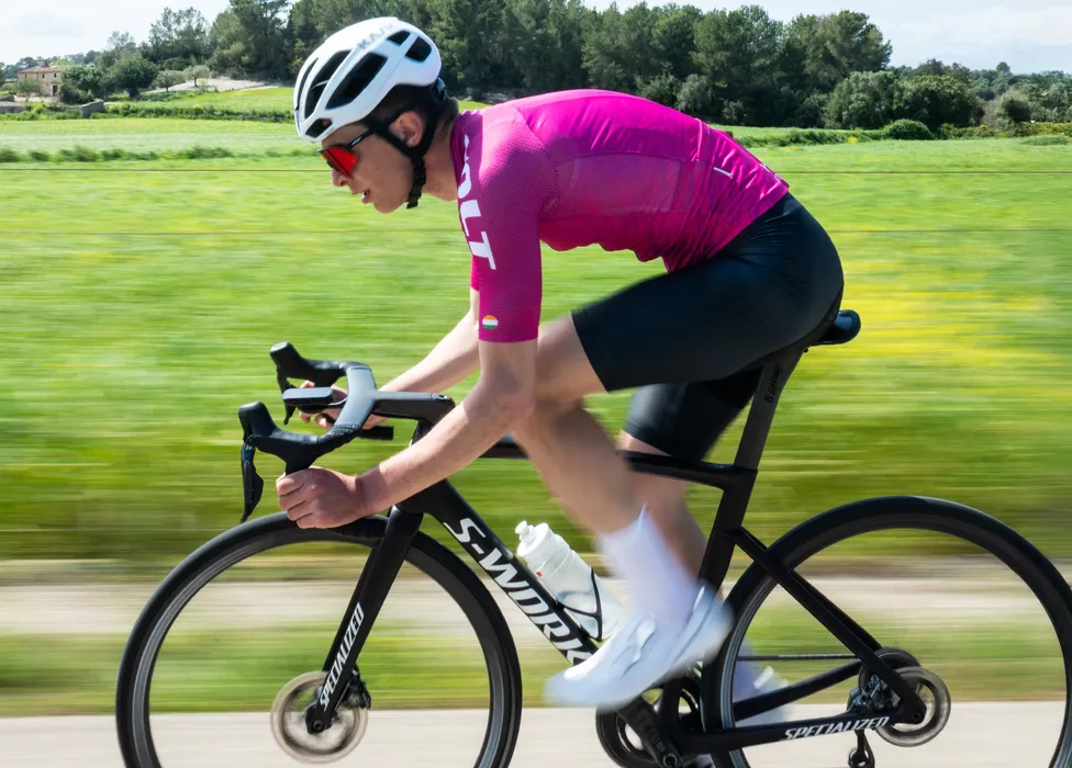 Un cycliste en maillot rose et short noir roule sur une route à travers un paysage verdoyant, la flou de mouvement soulignant la vitesse.