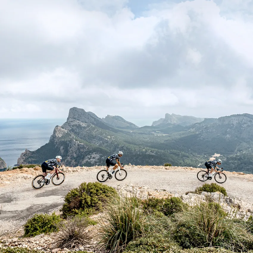 Trois cyclistes en tenue de sport roulent sur une route sinueuse avec un paysage montagneux et la mer en arrière-plan.