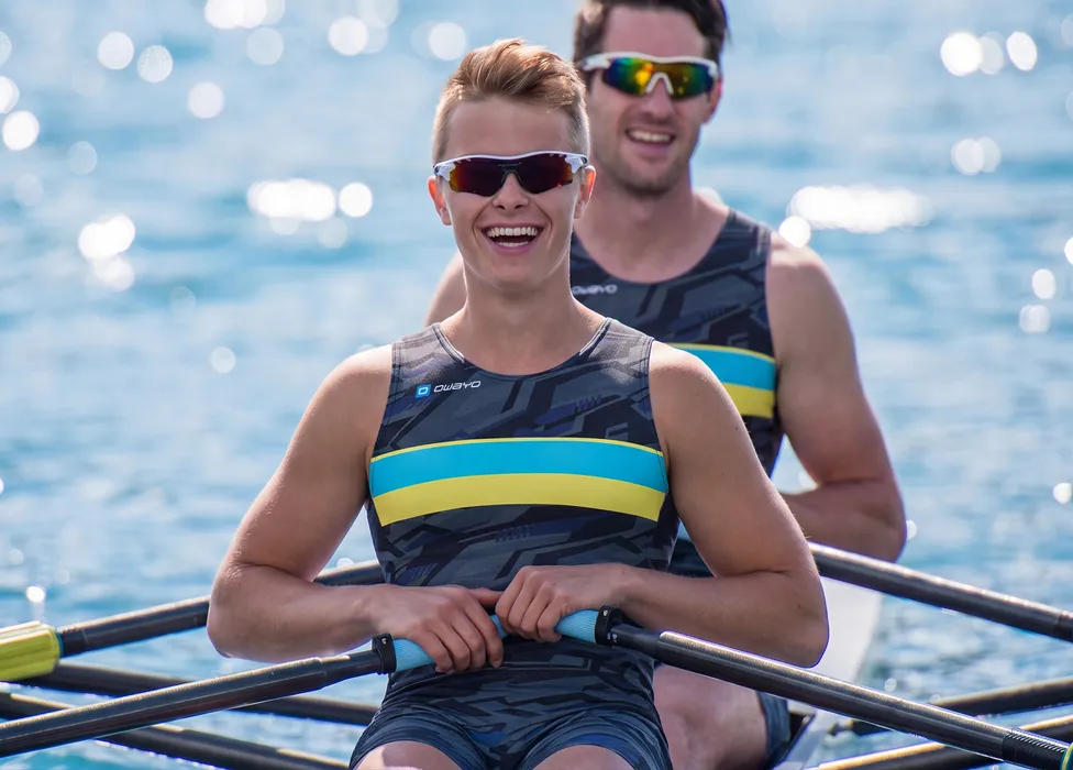 Deux rameurs dans des combinaisons sportives à motifs avec des rayures bleues et jaunes, souriant sur un plan d'eau.