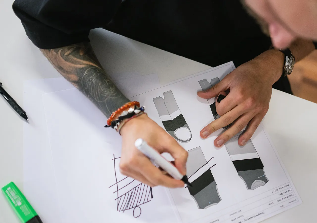 Une personne avec un tatouage sur le bras dessine avec un marqueur blanc sur une feuille de papier présentant plusieurs modèles de vêtements de sport.