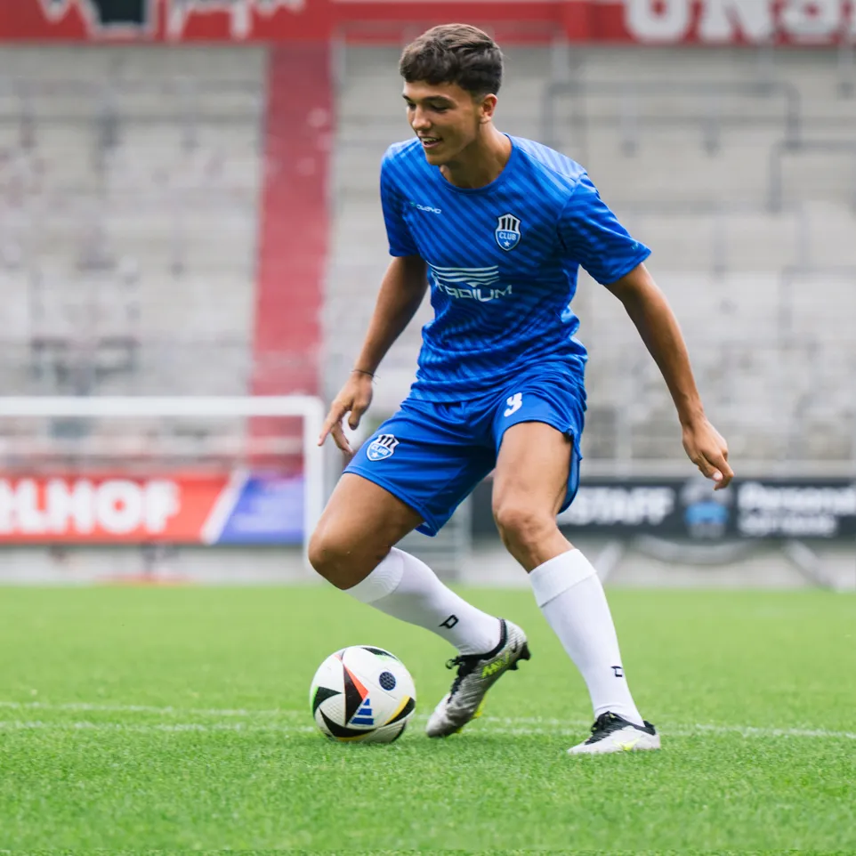 Un jeune joueur de football en maillot bleu et short bleu avec un logo sur la poitrine et le numéro 3 sur le short dribble un ballon de football sur un terrain d'herbe verte.