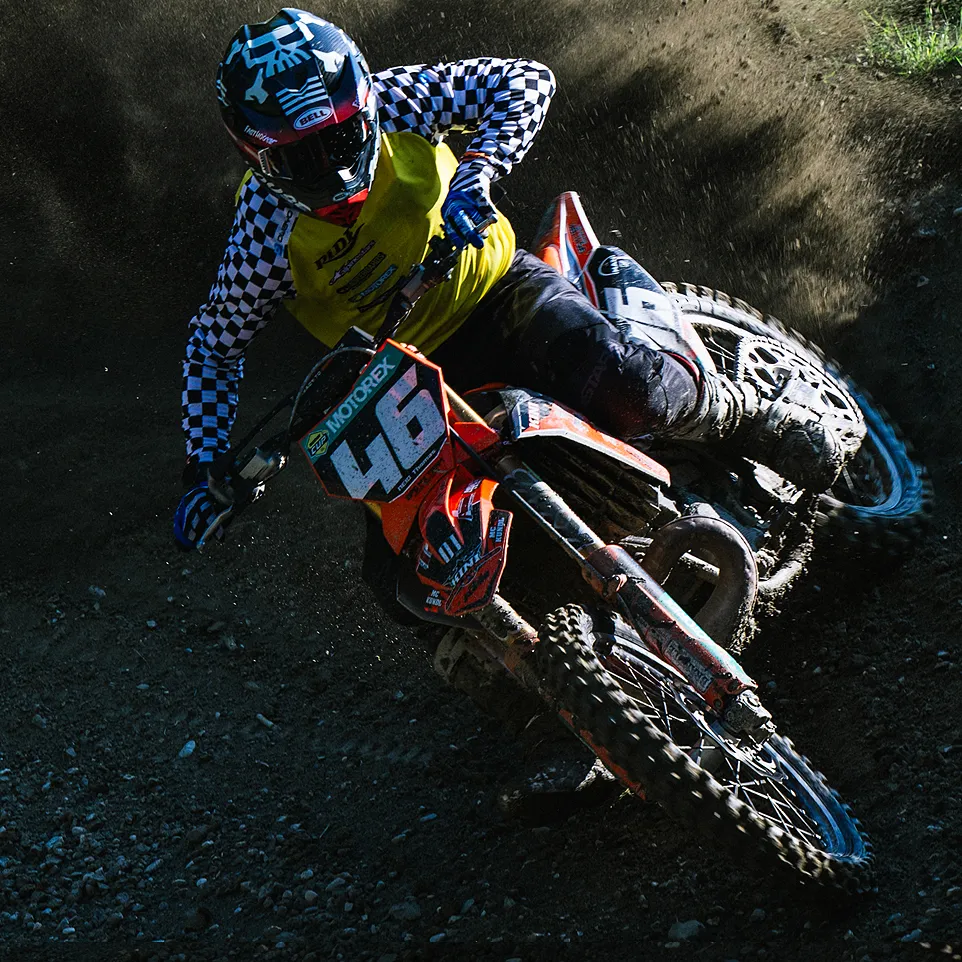 Un pilote de motocross en veste jaune avec des manches à carreaux noirs et blancs et un casque conduit une moto orange sur une piste poussiéreuse.