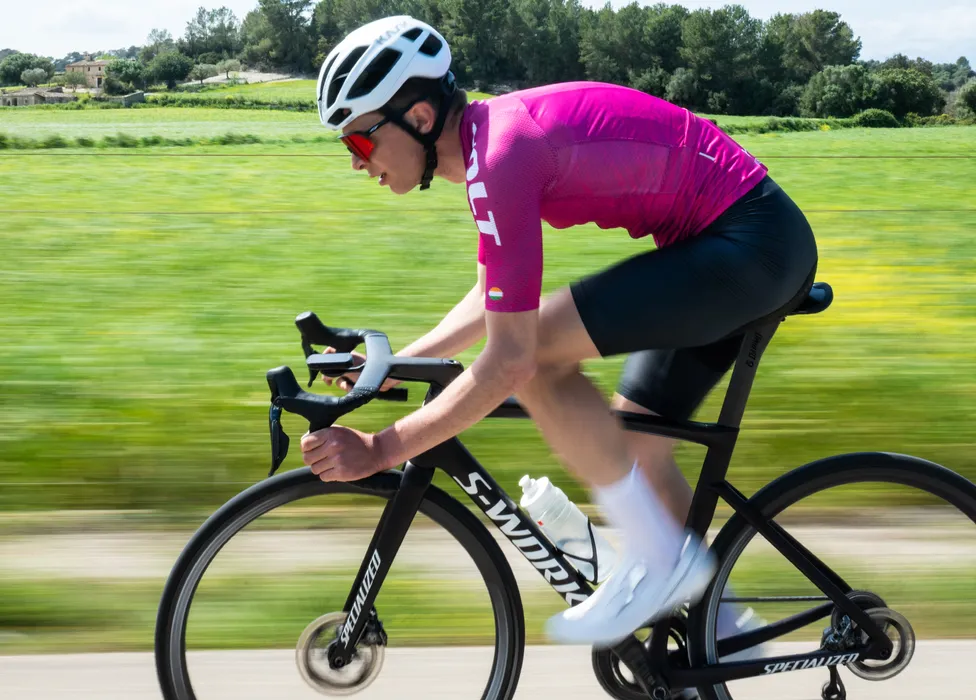 Un cycliste en maillot rose et short noir roule sur un vélo de route sur une route de campagne, avec un arrière-plan vert flou suggérant de la vitesse.