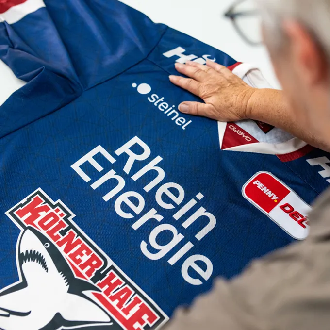 Une personne examine un maillot sportif bleu personnalisé d'owayo avec différents logos de sponsors, notamment 'Rhein Energie' et 'Kölner Haie'.
