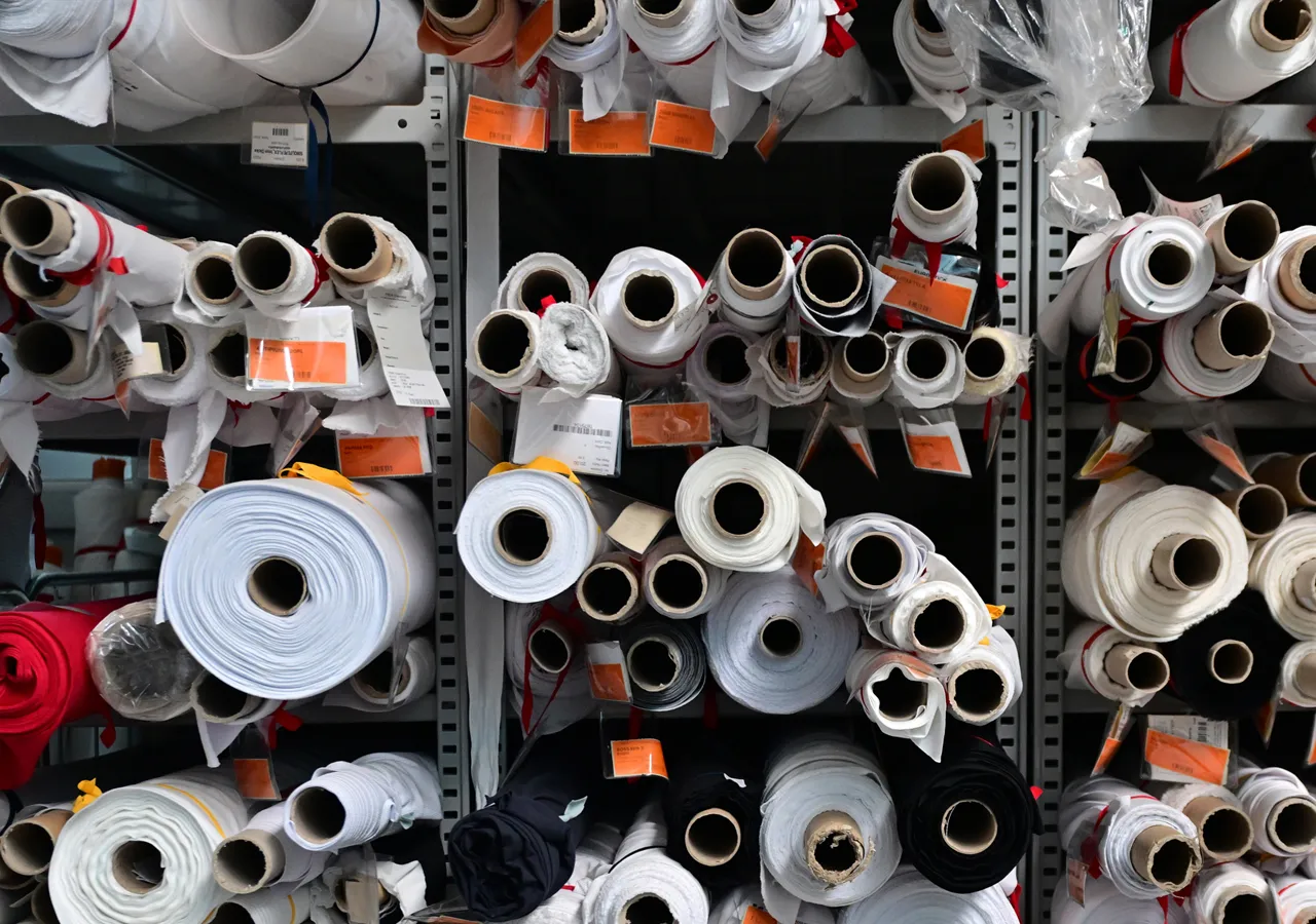 Des étagères remplies de rouleaux de tissu de différentes couleurs, notamment blanc, noir et rouge, avec des étiquettes indiquant une usine de production interne.