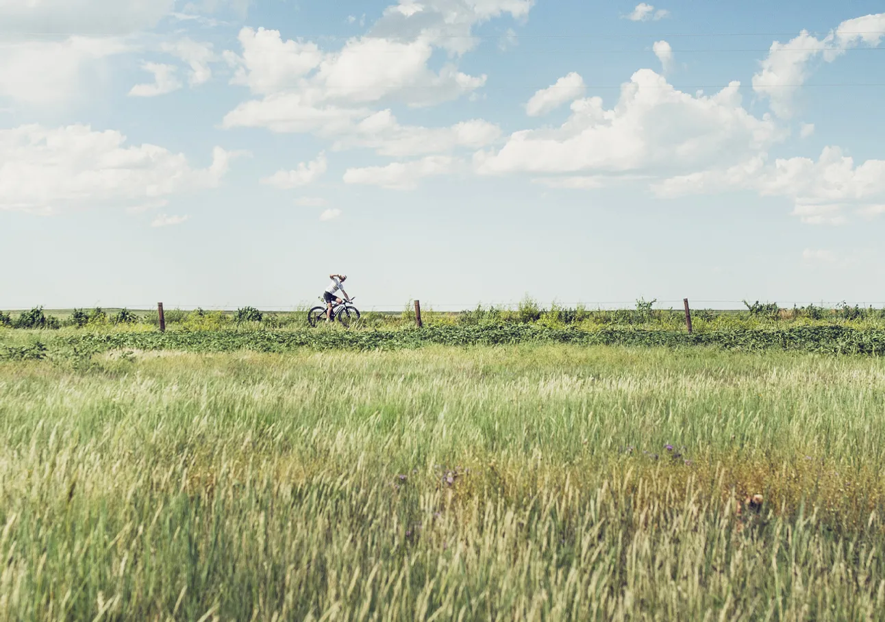 Une personne fait du vélo dans un champ vert sous un ciel bleu avec des nuages blancs.